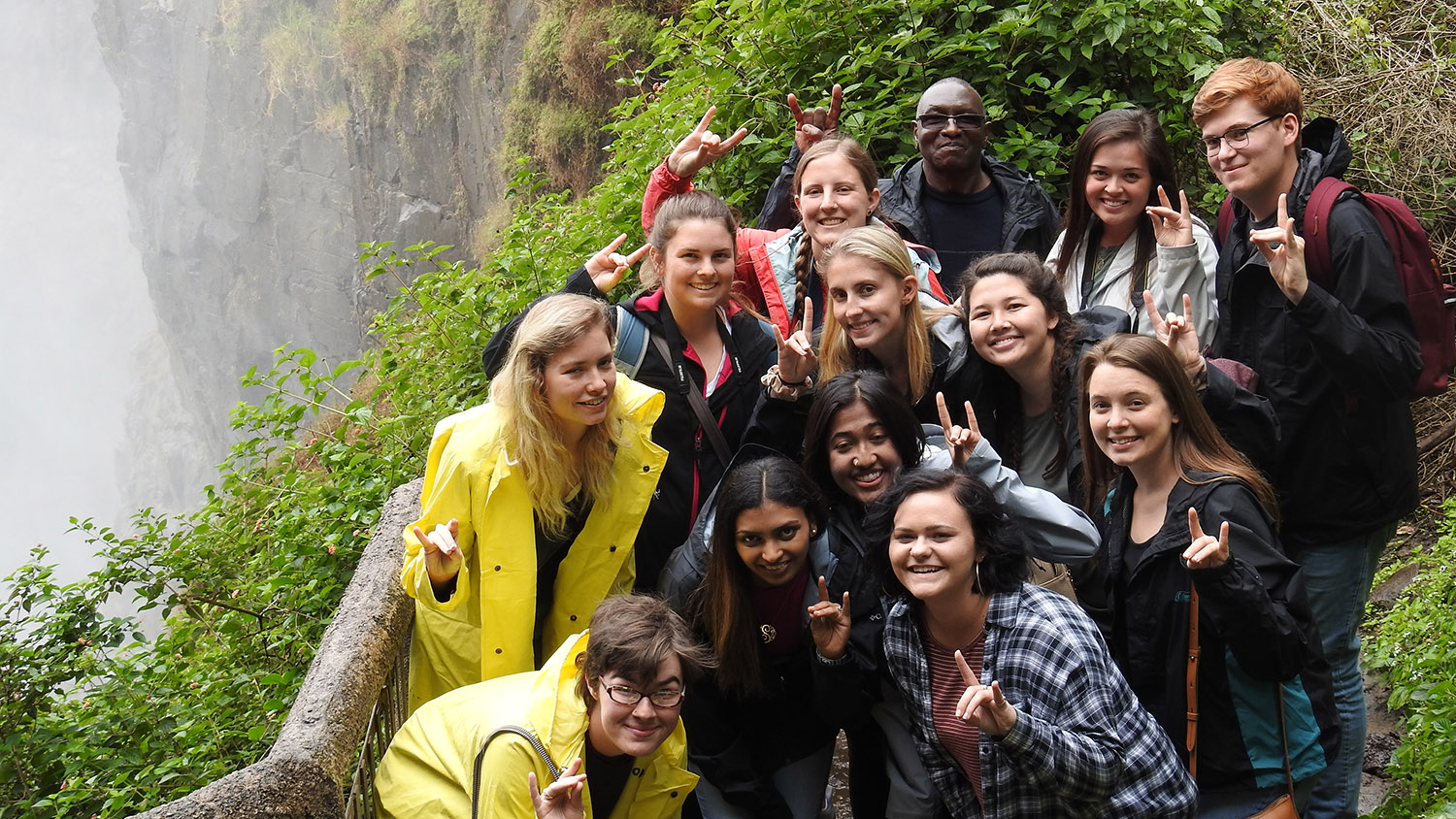 students hold up "wolfie" hand sign while standing near waterfall